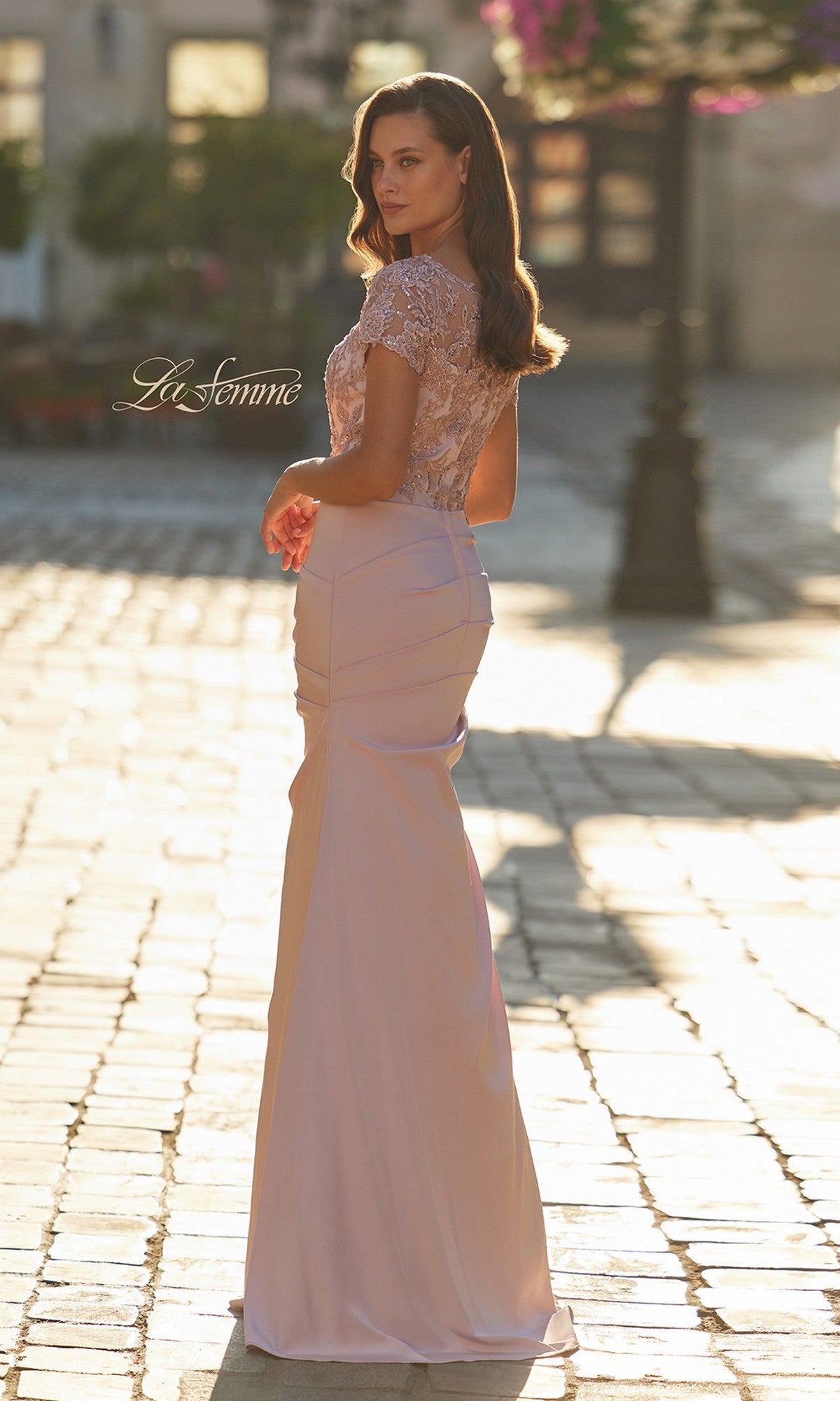 A woman stands outdoors on a sunlit cobblestone path, wearing the La Femme 32737 long formal dress in light pink. This gown features an illusion neckline, lace-embroidered bodice, and elegant cap sleeves.